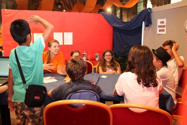 Eine Gruppe von Kindern sitzt an einem Tisch, während eine Frau mit ihnen spricht.