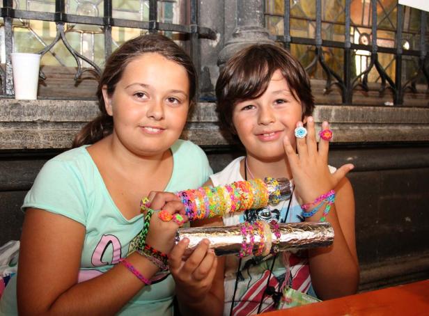Zwei Kinder präsentieren selbstgemachten Schmuck aus bunten Gummibändern.