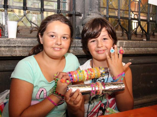 Zwei Kinder präsentieren selbstgemachten Schmuck aus bunten Gummibändern.