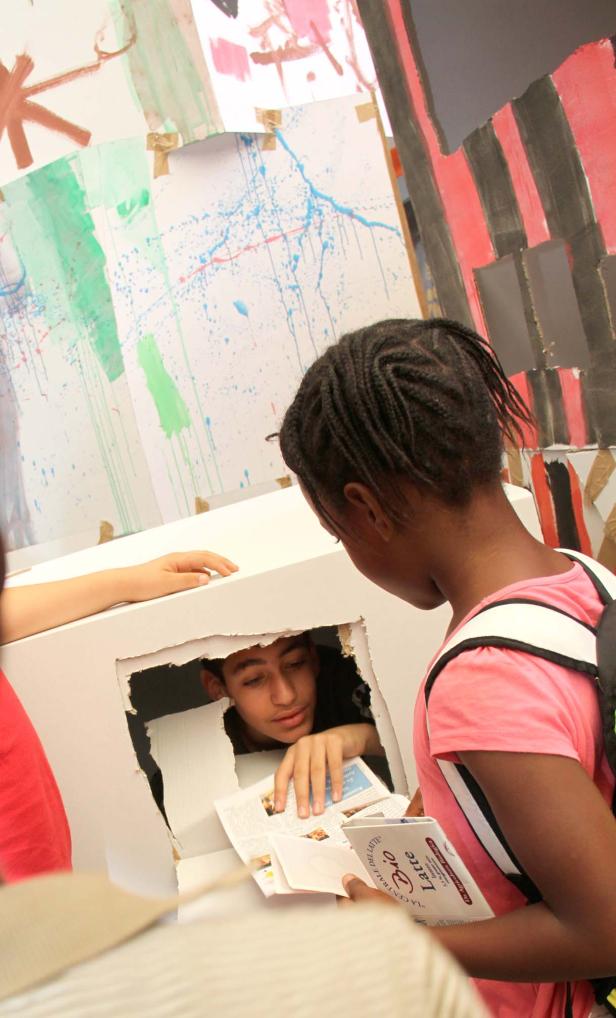 Ein Junge schaut aus einem Loch in einer bemalten Pappwand, während ein Mädchen ihm Papiere reicht.