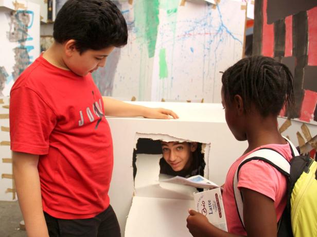 Zwei Kinder stehen vor einem Karton, aus dessen Öffnung ein Jugendlicher herausschaut.