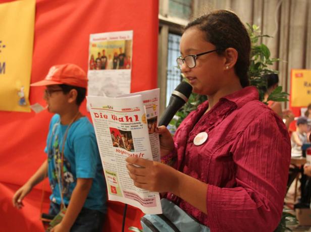 Ein junges Mädchen liest mit einem Mikrofon in der Hand aus einer Zeitung mit dem Titel „Die Wahl“ vor.