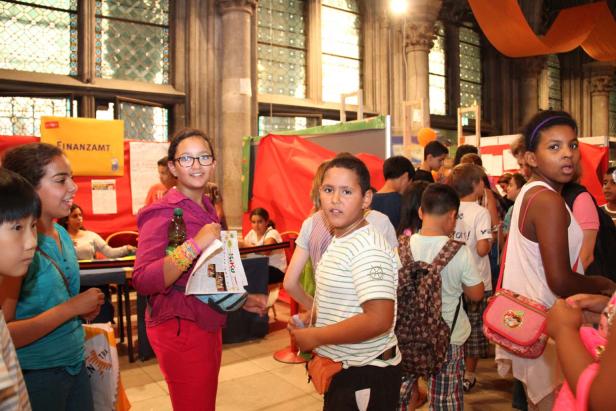 Eine Gruppe von Kindern steht in einer Halle mit Ständen und bunten Dekorationen.