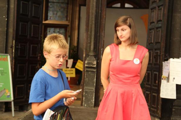 Ein Junge schaut auf sein Handy, während eine Frau in einem roten Kleid daneben steht.