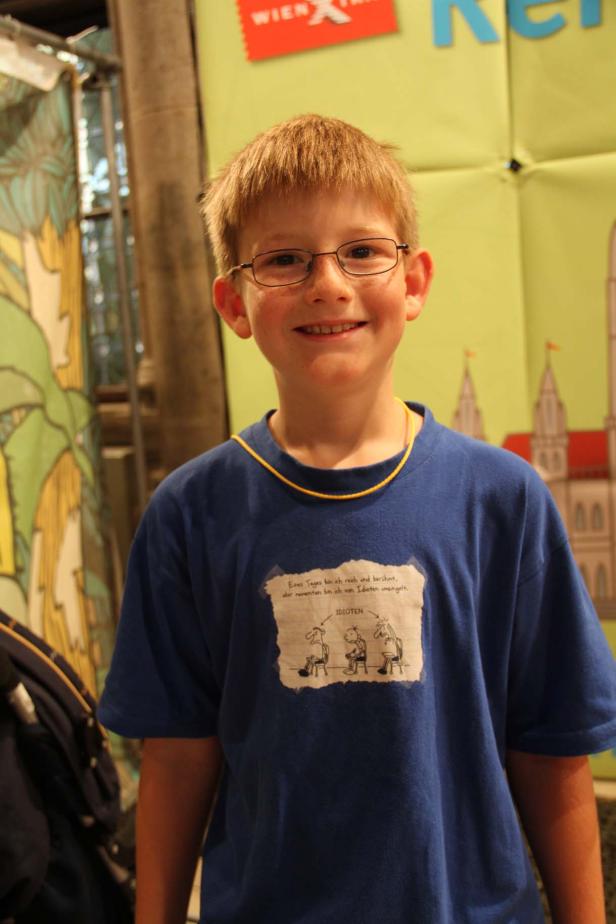 Ein lächelnder Junge mit Brille und blauem T-Shirt steht vor einem bunten Hintergrund.