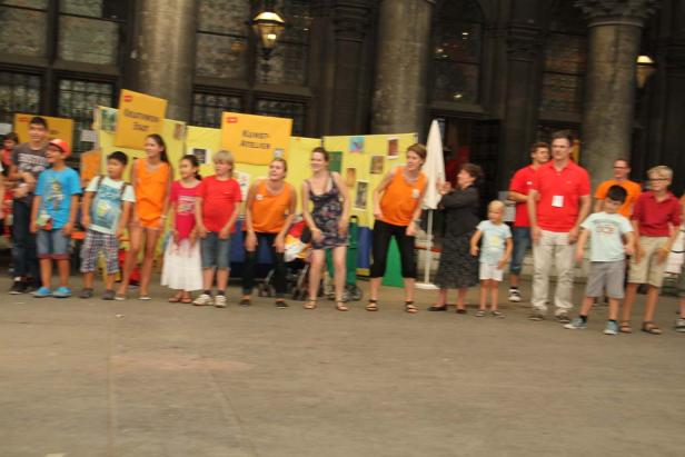 Eine Gruppe von Kindern und Erwachsenen steht vor einem Stand mit gelben Plakaten.
