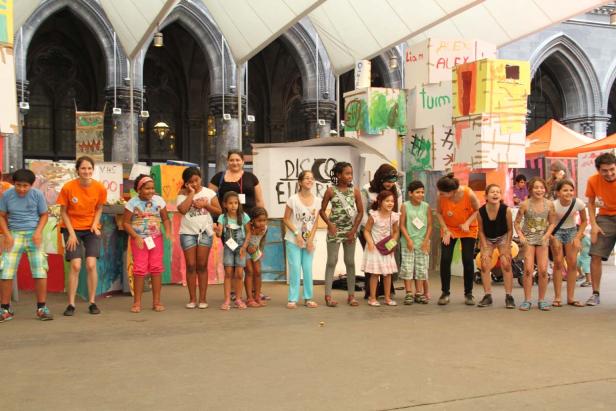 Eine Gruppe Kinder und Erwachsene posiert vor bemalten Kartons und einem Gebäude mit Bögen.