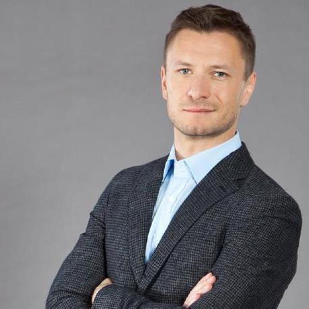 Ein Mann mit blauem Hemd und Sakko vor einem grauen Hintergrund.