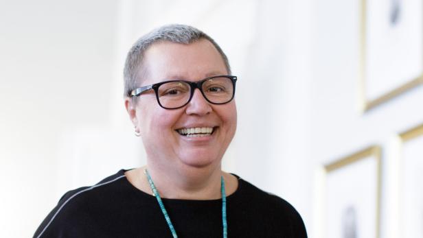 Eine lächelnde Frau mit Brille und kurzem, grauem Haar trägt eine türkisfarbene Halskette.