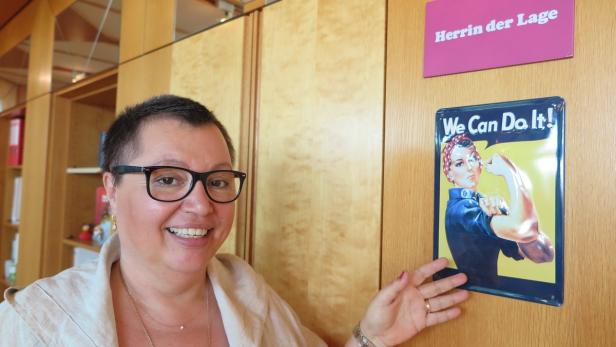 Eine Frau steht neben einem „We Can Do It!“-Poster und einem Schild mit der Aufschrift „Herrin der Lage“.