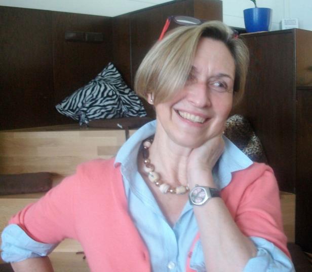 Eine lächelnde Frau mit kurzem blonden Haar, Perlenkette und korallfarbener Strickjacke posiert für ein Foto.