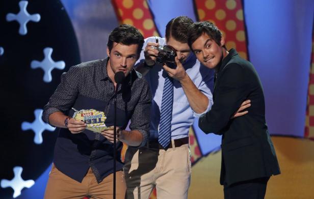 Drei Männer posieren auf einer Bühne, einer mit Mikrofon und „Teen Choice“-Award, einer mit Kamera.