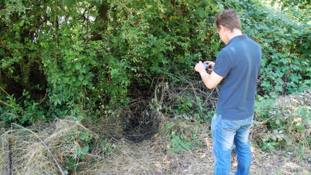 Ein Mann fotografiert mit seinem Smartphone ein Loch im Gebüsch.