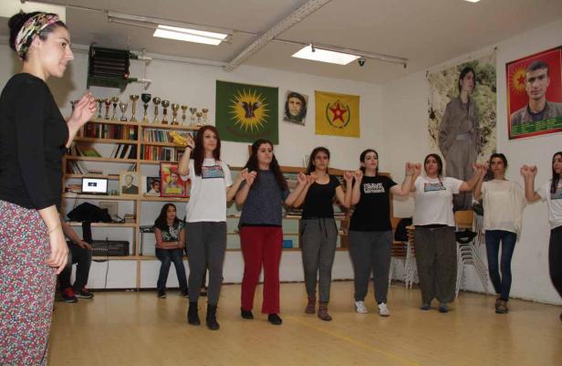 Eine Gruppe junger Frauen tanzt in einem Raum mit kurdischen Flaggen und Porträts.