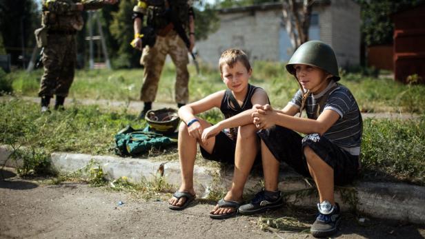 Zwei Jungen sitzen am Straßenrand, im Hintergrund Soldaten.