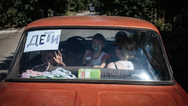 Eine Familie flieht in einem Auto, an dessen Heckscheibe ein Schild mit der Aufschrift „Kinder“ befestigt ist.