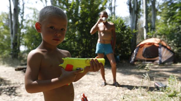 Ein Junge zielt mit einer gelben Spielzeugpistole, im Hintergrund ein Mann und ein Zelt.