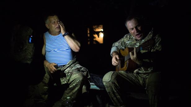 Zwei Männer sitzen im Dunkeln, einer singt und spielt Gitarre, der andere wischt sich eine Träne aus dem Auge.