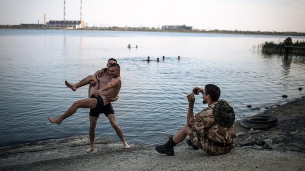 Ein Mann wird von einem anderen am Ufer eines Sees getragen, während ein Dritter ein Foto macht.