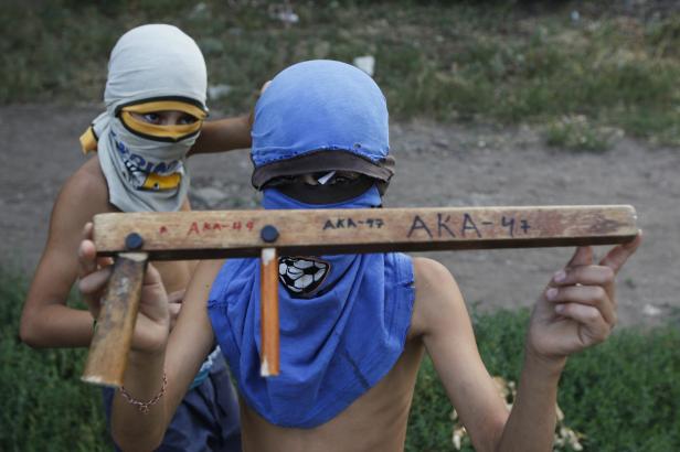 Zwei Kinder mit improvisierten Masken spielen mit einem Holzgewehr, auf dem „AKA-47“ steht.
