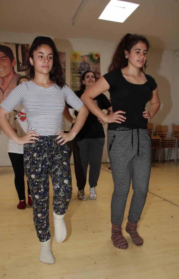 Zwei junge Frauen tanzen in einem Raum mit anderen Personen und Postern an der Wand.