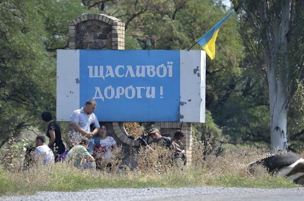 Eine Gruppe von Menschen steht vor einem Schild mit der Aufschrift „Щасливої дороги!“ und einer ukrainischen Flagge.