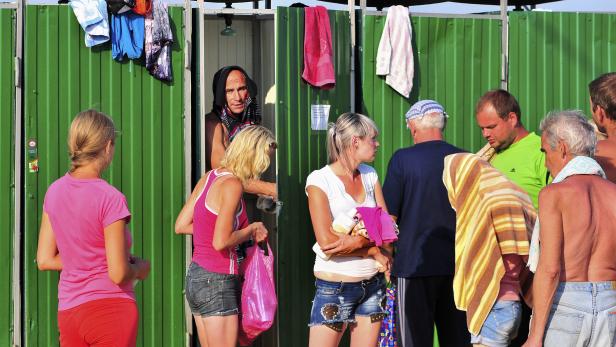 Menschen stehen vor grünen Umkleidekabinen am Strand.