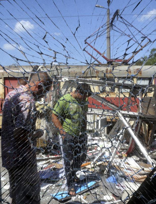 Zwei Männer stehen vor einer zerstörten Fassade, gesehen durch eine zerbrochene Scheibe.