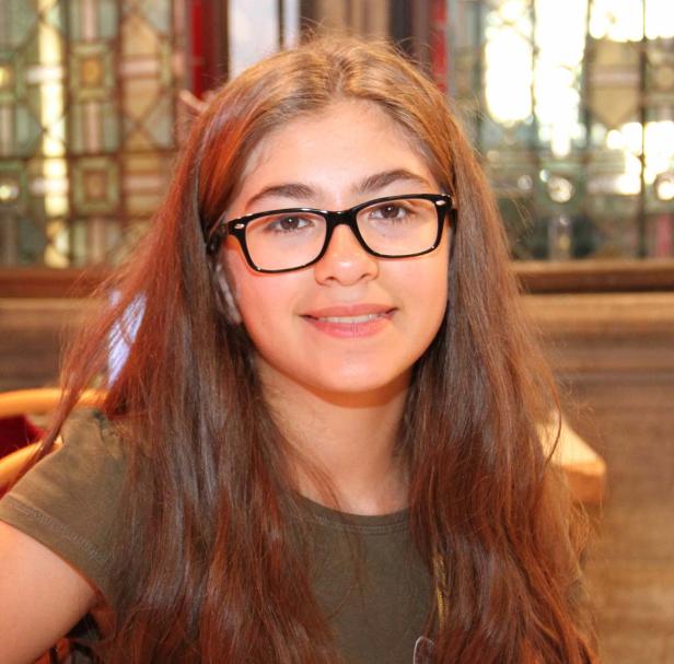 Ein junges Mädchen mit braunen Haaren und Brille lächelt in die Kamera.