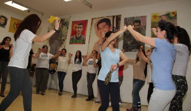 Eine Gruppe von Frauen tanzt in einem Raum mit kurdischen Flaggen und Porträts an der Wand.