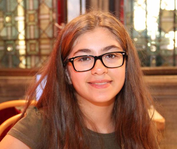 Ein junges Mädchen mit braunen Haaren und Brille lächelt in die Kamera.