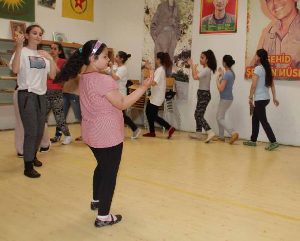 Eine Gruppe junger Frauen tanzt in einem Raum mit kurdischen Flaggen und Porträts an der Wand.