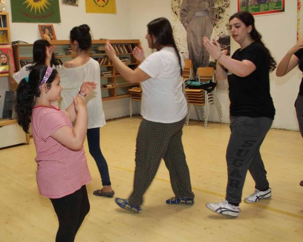Eine Gruppe von Frauen und ein Mädchen tanzen in einem Raum mit kurdischen Flaggen.