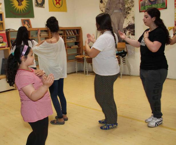 Eine Gruppe von Frauen und ein Mädchen tanzen in einem Raum mit kurdischen Flaggen.