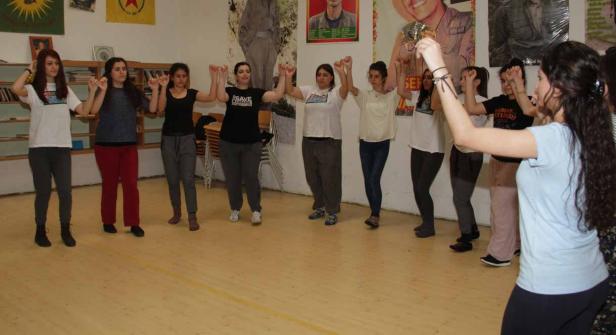 Eine Gruppe junger Frauen tanzt im Kreis in einem Raum mit kurdischen Flaggen und Postern.