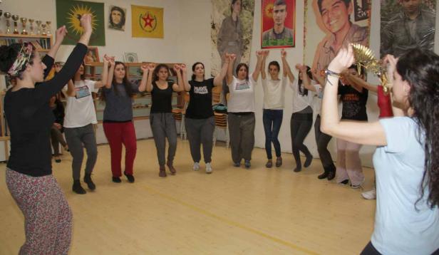 Eine Gruppe junger Frauen tanzt in einem Raum mit kurdischen Flaggen und Porträts an der Wand.