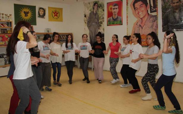 Eine Gruppe junger Frauen tanzt im Kreis in einem Raum mit kurdischen Flaggen und Porträts an den Wänden.