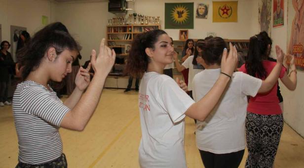 Eine Gruppe junger Frauen tanzt in einem Raum mit kurdischen Flaggen und Postern an der Wand.