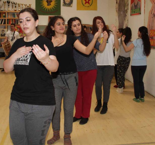 Eine Gruppe junger Frauen tanzt in einem Raum mit kurdischen Flaggen an der Wand.