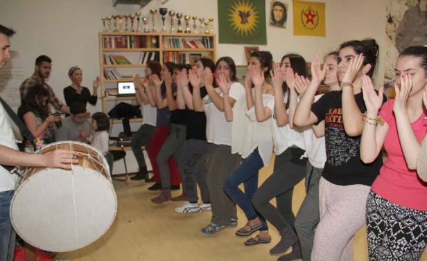 Eine Gruppe von Frauen tanzt zu den Klängen einer Davul-Trommel in einem Raum mit kurdischen Flaggen.