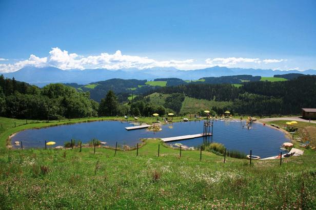 Ein Badesee mit Liegewiese und Sprungturm vor einer bewaldeten Hügellandschaft.