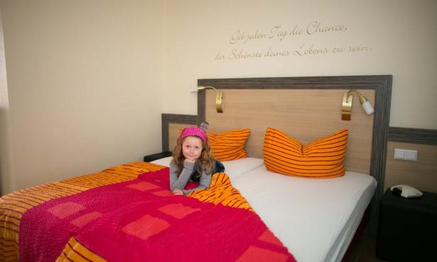 Ein Mädchen liegt auf einem Bett mit orangefarbenen Kissen in einem Hotelzimmer.