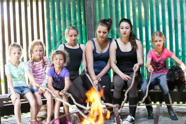 Eine Gruppe von Mädchen und eine junge Frau sitzen mit einem Hund an einem Lagerfeuer.