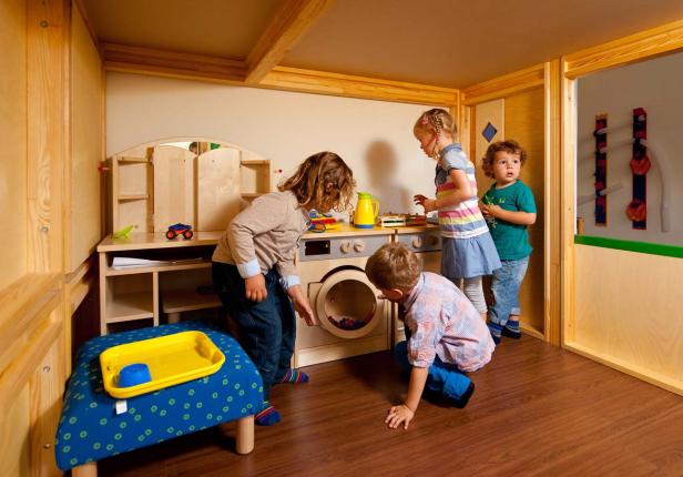 Vier Kinder spielen in einem hölzernen Spielzimmer mit Spielküche und Waschmaschine.