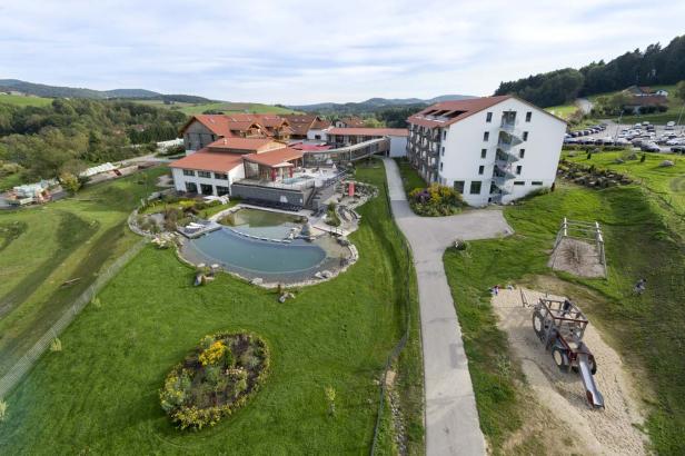 Luftaufnahme eines Hotels mit Schwimmteich und Spielplatz in einer grünen Landschaft.