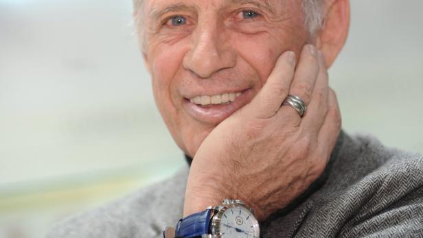 Ein älterer Mann mit einem grauen Jackett und einer blauen Uhr lächelt in die Kamera.