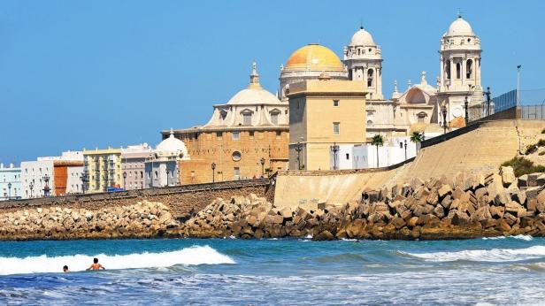 Blick auf die Kathedrale von Cádiz und die Küstenlinie mit Badegästen im Wasser.