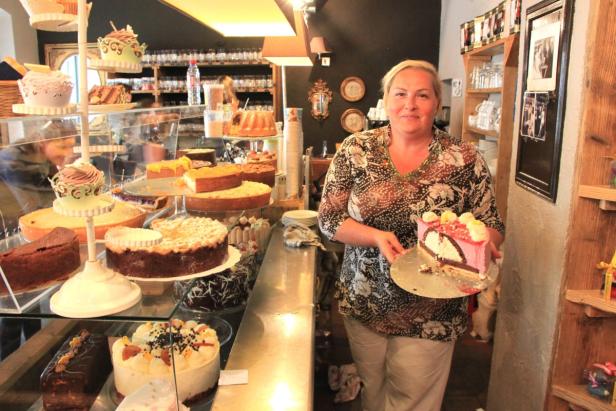 Eine Frau präsentiert ein Stück rosa Torte in einem Café mit vielen Kuchen.