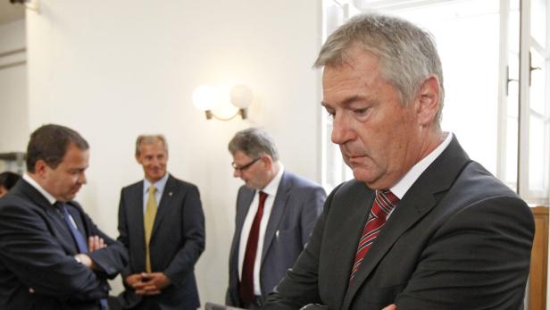 Ein Mann im Anzug steht mit verschränkten Armen vor einer Gruppe von Personen.