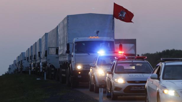 Ein Konvoi von Lastwagen wird von Polizeiautos mit Blaulicht begleitet; eine rote Flagge weht im Wind.